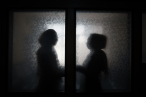 A silhouette of two female figures in modest dress and headscarves, stood in front of patterned glass windows or doors.